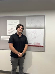 Noah White stands beside a display of research and design materials mounted on a wall.