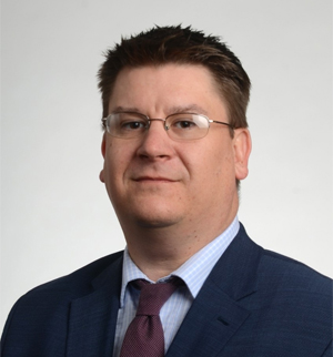 Headshot of Dean Jim Buss, a dark haired man with glasses in a dark blue suit and purple tie. 