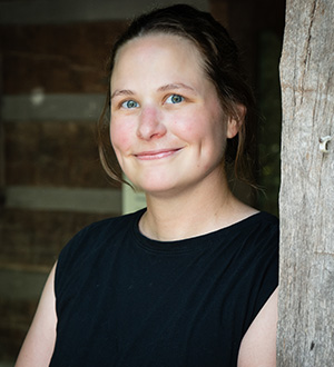Woman smiling outdoors while standing beside a wooden post, wearing a black sleeveless top.