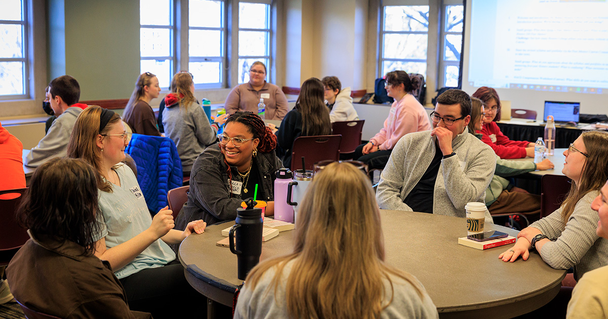 Students sit, talk, and laugh with each other at a circular table in a well-lit classroom