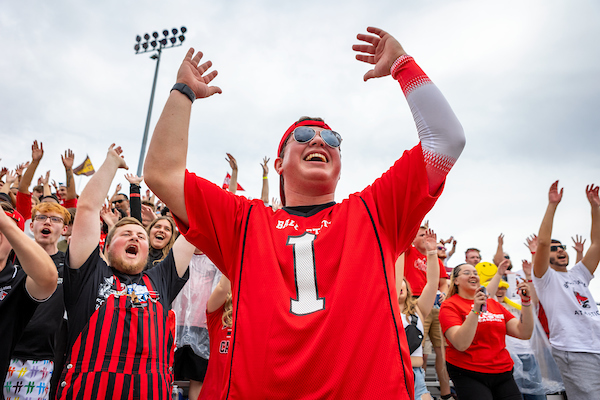 A low camera view shows a crowd of fans dressed in red and white BSU gear cheering against a bright but cloudy sky.