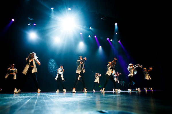 A group dressed in black with open white shirts dances on a stage with bright blue lights over their heads