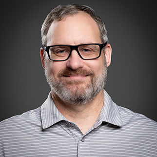 A man wearing glasses and a grey and white, striped button up shirt in front of a gray background