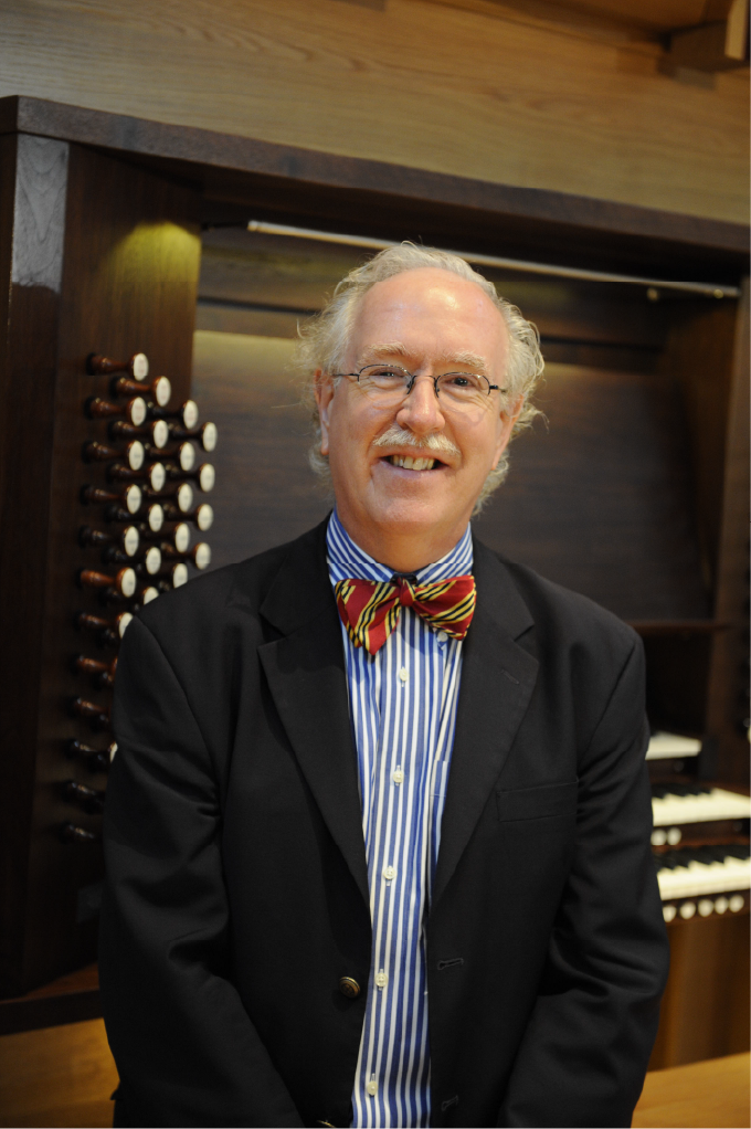 Colin Andrews, organist, wears a black suit, blue striped shirt, and bowtie standing before an organ.