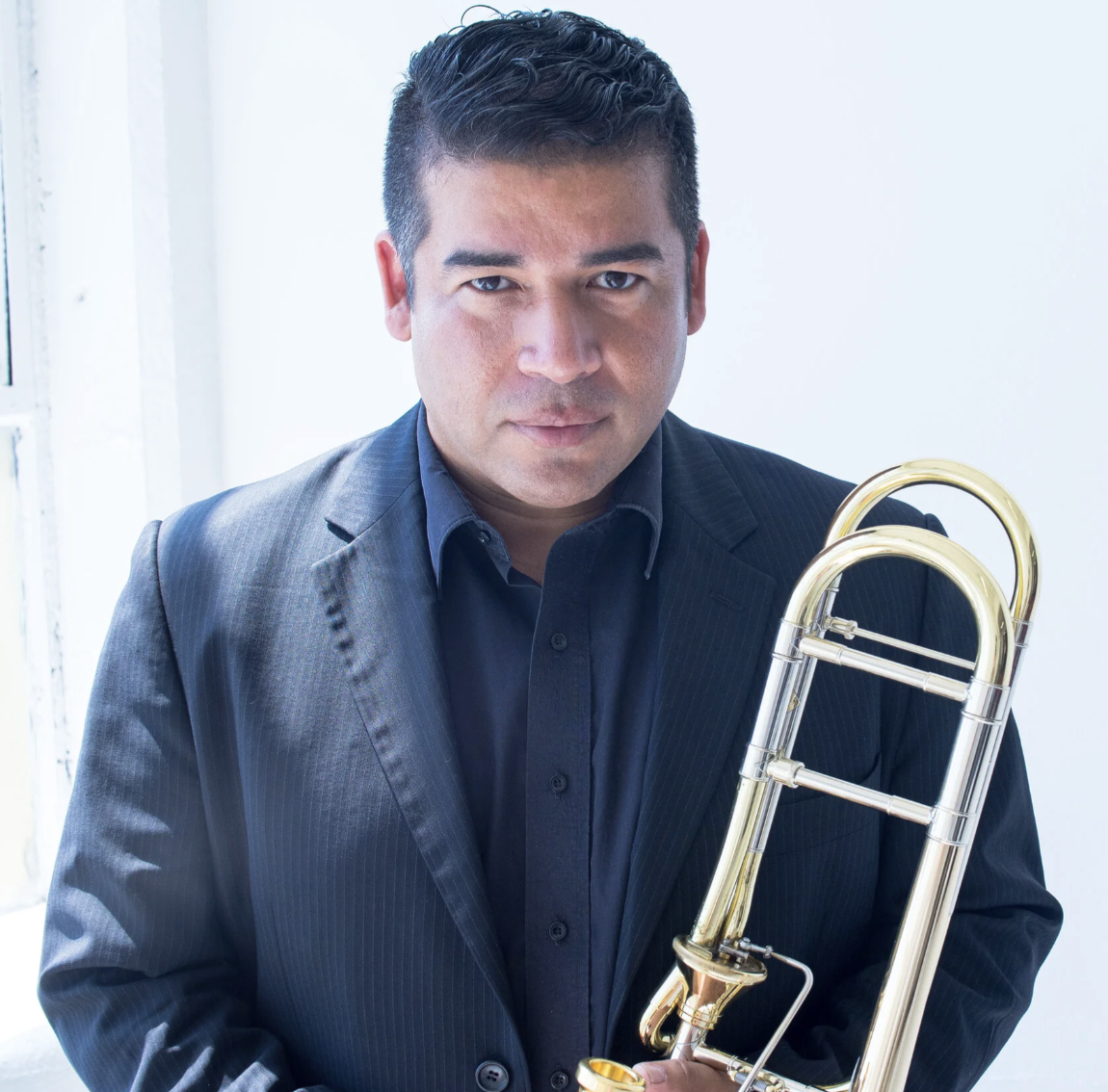 Dr. Denis Jiron looks at the camera holding a trombone while wearing a navy suit.