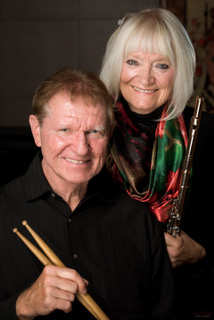 John and Paula Kasica pose together with John holding a pair of drumsticks and Paula holding a flute.