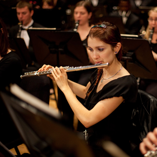 A flute player looking down at a music stand with other woodwind players in the background