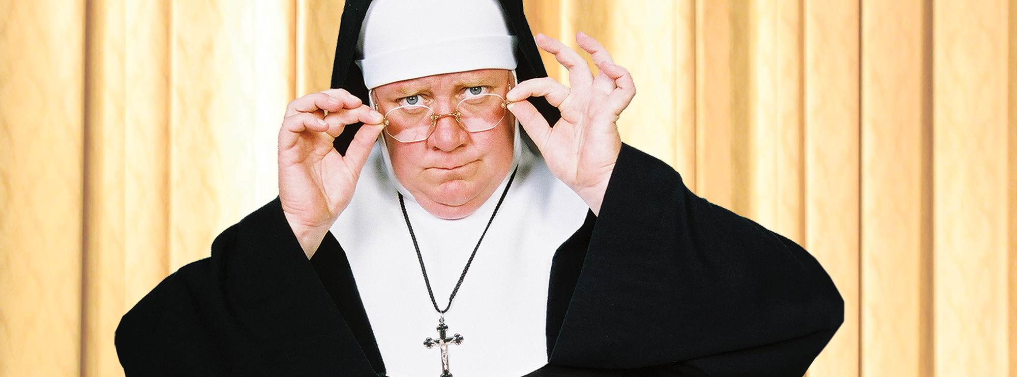 A person is dressed in a nun costume with cross necklace, holding their glasses in judgement in front of a khaki curtain.