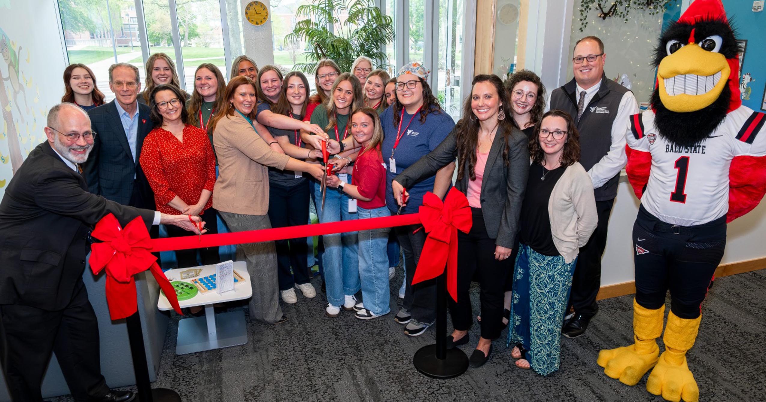 Group of Ball State students, faculty, and community partners cut a red ribbon to celebrate the newly redesigned Interprofessional Community Clinics waiting area, with the Ball State mascot standing nearby.