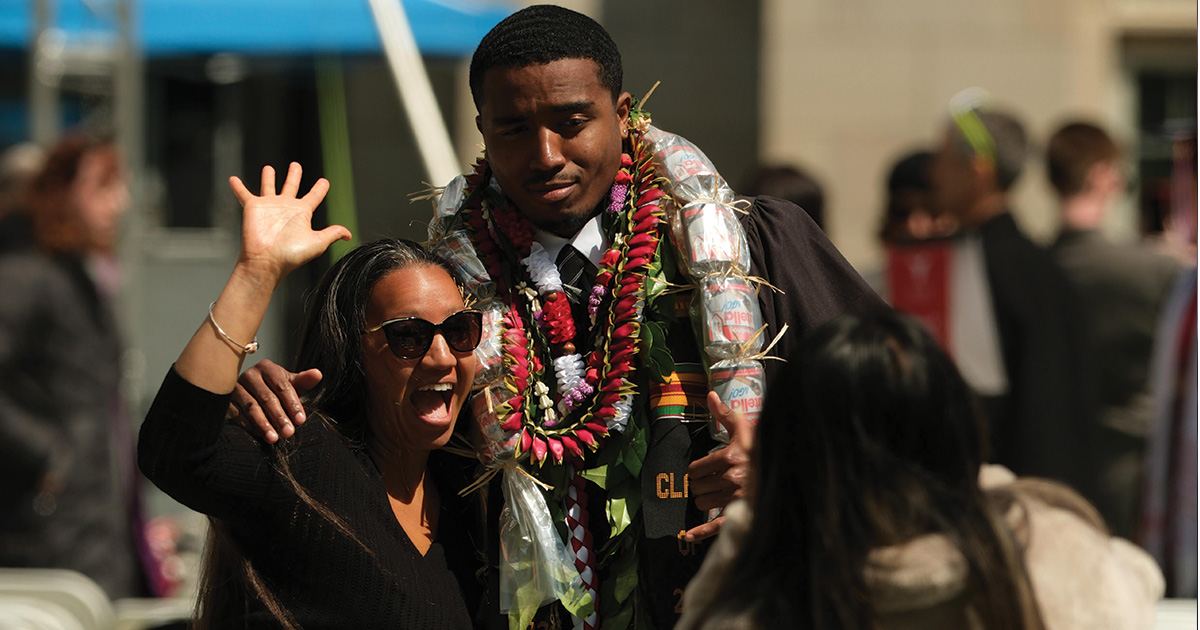 Graduate poses with arm around mother who is smiling and waving.