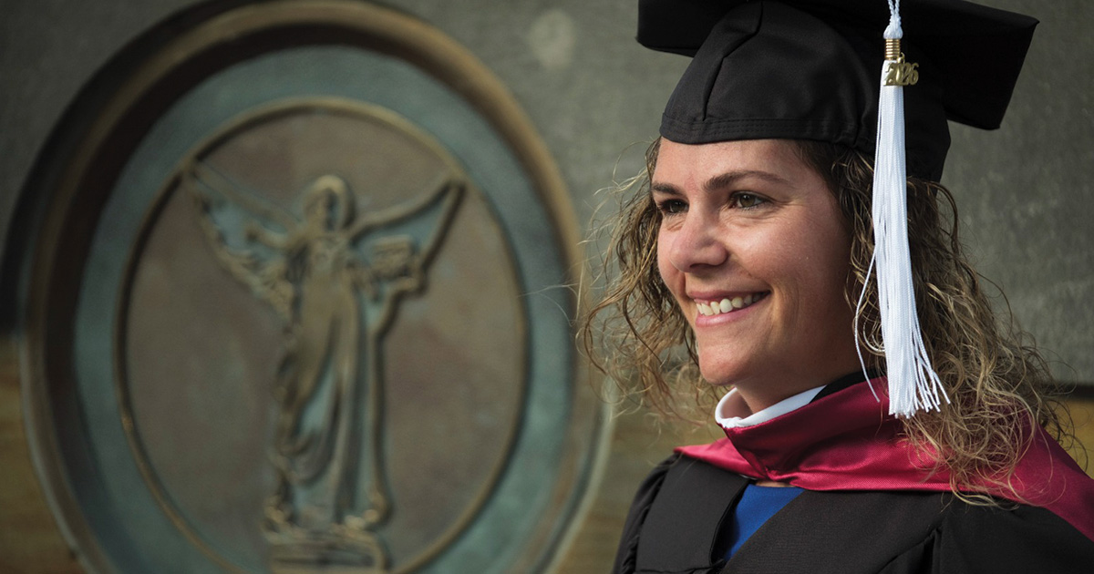 Graduate poses in cap and gown beside the Beneficence medallion.