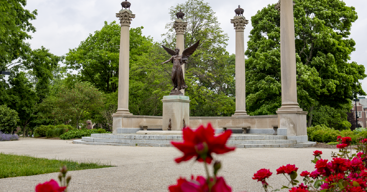 Beneficence statue on Ball State's campus with trees in the background and red flowers in front.